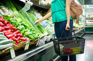 Adult with brown bag and wearing green shirt and blue jeans with black market basket choosing produce from baskets of capsicums and shallots in supermarket.